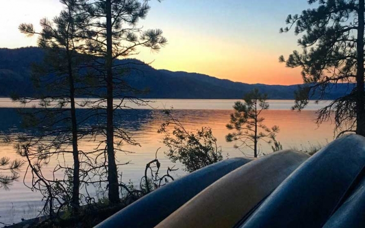 a group of canoes rest on the shore beside a calm lake while the sun is setting